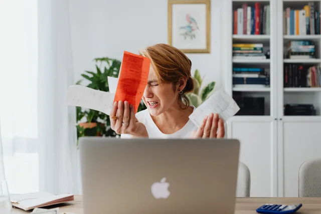 Frustrated woman managing finances, surrounded by bills and a laptop, looking distressed. Kassekredit eller likviditetsbuffer: hvad koster det at stå klar?